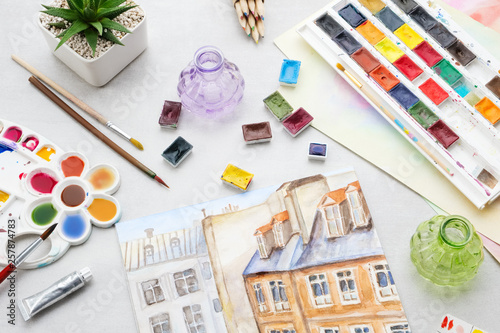 Watercolor drawing - view of the roofs of houses. Artistic equipment on desk. Top view.