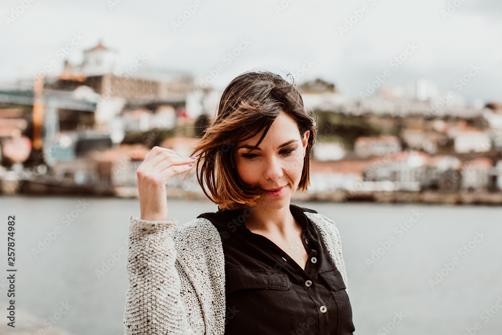 .Short haired pretty woman sightseeing in the city of Porto, Portugal. Standing on the river side, enjoying a beautiful landscape on a windy spring day. Lifestyle.