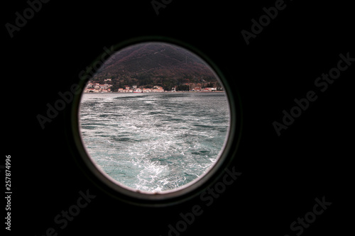 Hinged round window  storm cover on ship looking outside into Mediterranean sea. Porthole view through window on ship to turbulence made by foam of sea water from a high-speed boat on surface of sea.