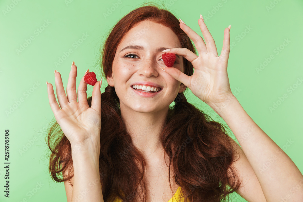 Obraz premium Emotional young redhead girl posing isolated over green wall background with raspberry.