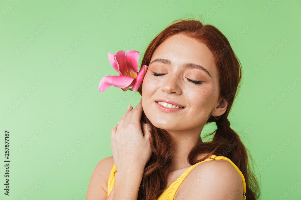 Happy young redhead girl posing isolated over green wall background holding flower. Happy young redhead girl posing isolated over green wall background holding flower.
