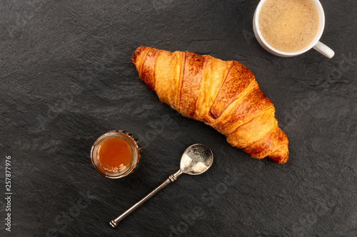 A closeup photo of a croissant with a cup of coffee and a jar of jam on a black background with a place for text