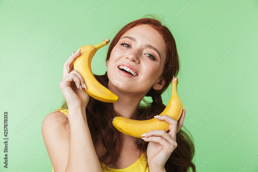 Excited young redhead girl posing isolated over green wall background with bananas fruits. Excited young redhead girl posing isolated over green wall background with bananas fruits.