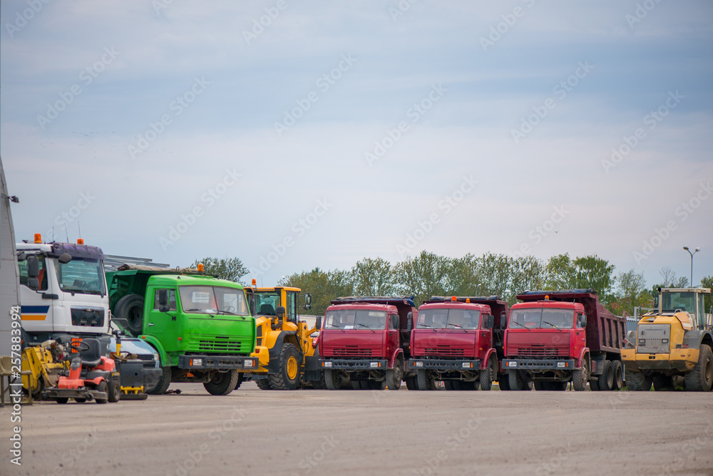 Multiple cars, trucks, loaders, concrete mixers and construction machinery in large parking lot in industrial territory, next to concrete and asphalt factory   