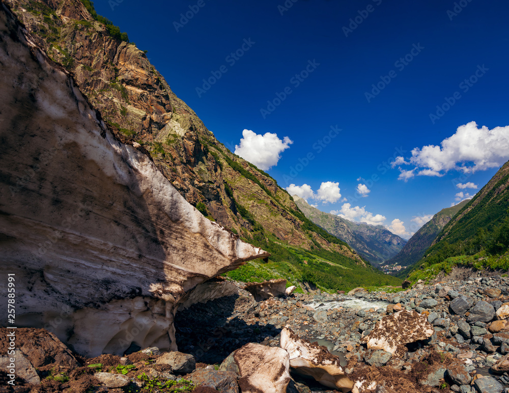 A huge block of snow melts in the summer in the Caucasian mountains near Dombai and a stream flows nearby.