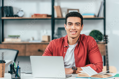 Smiling student in red shirt sitting at table and using laptop photo