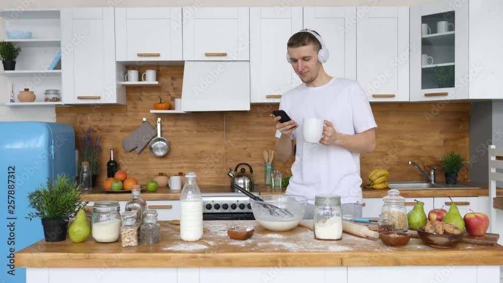 Young Man Using Mobile Phone In Kitchen At Home Wearing Headphones Young Man Using Mobile Phone In Kitchen At Home Wearing Headphones