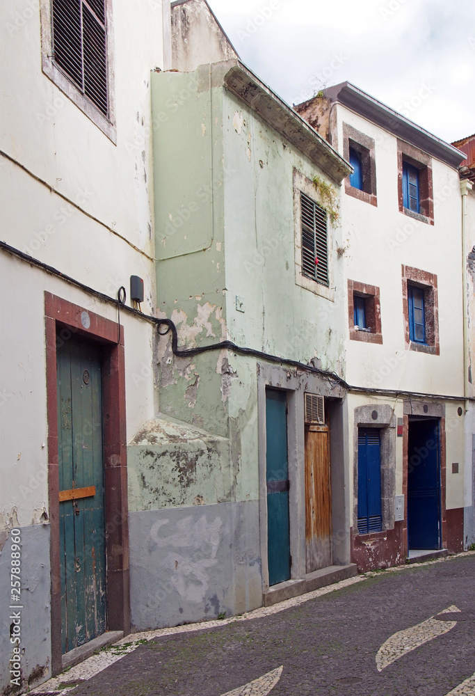 a typical quiet empty street in funchal madeira with old traditional houses painted in faded pastel peeling paint and a cobbled road