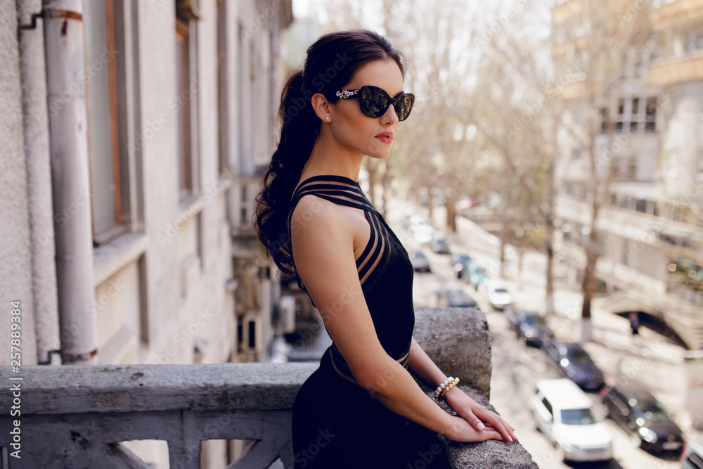 Strong, elegant woman in black sunglasses,sexy black dress, hair ponytail,  looks with attitude at the balcony. Stock Photo | Adobe Stock