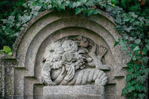 Mit Efeu bewachsener Grabstein mit einem Relief mit Blumen, Vögeln und Tauben als Grabschmuck auf dem Friedhof in Berlin Frohnau