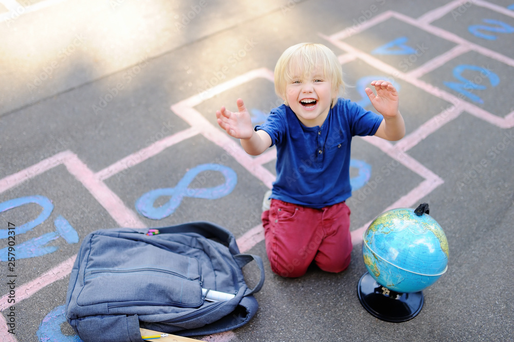 Naklejka premium Cute blond boy playing hopscotch game after school with bags laying near