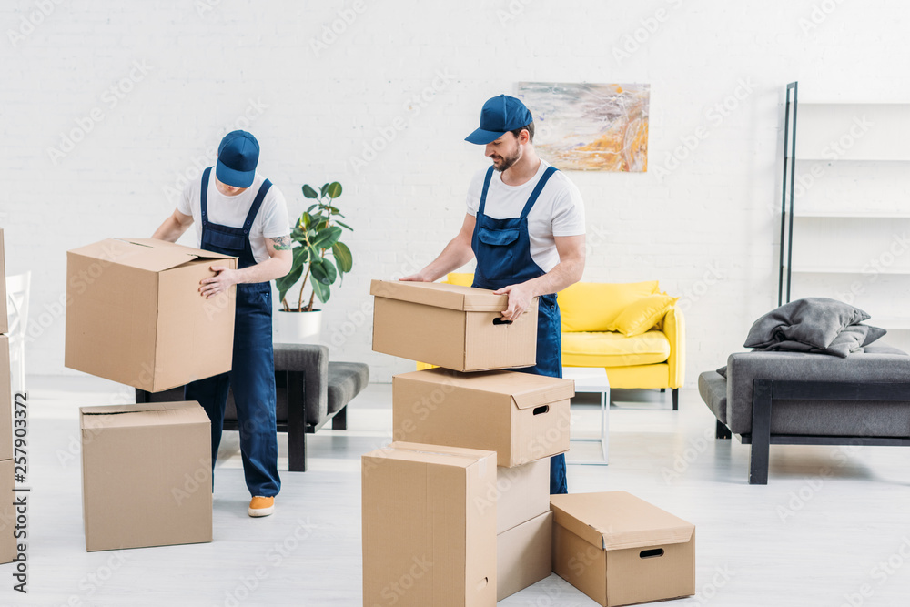 two movers in uniform carrying cardboard boxes in modern apartment - obrazy, fototapety, plakaty 