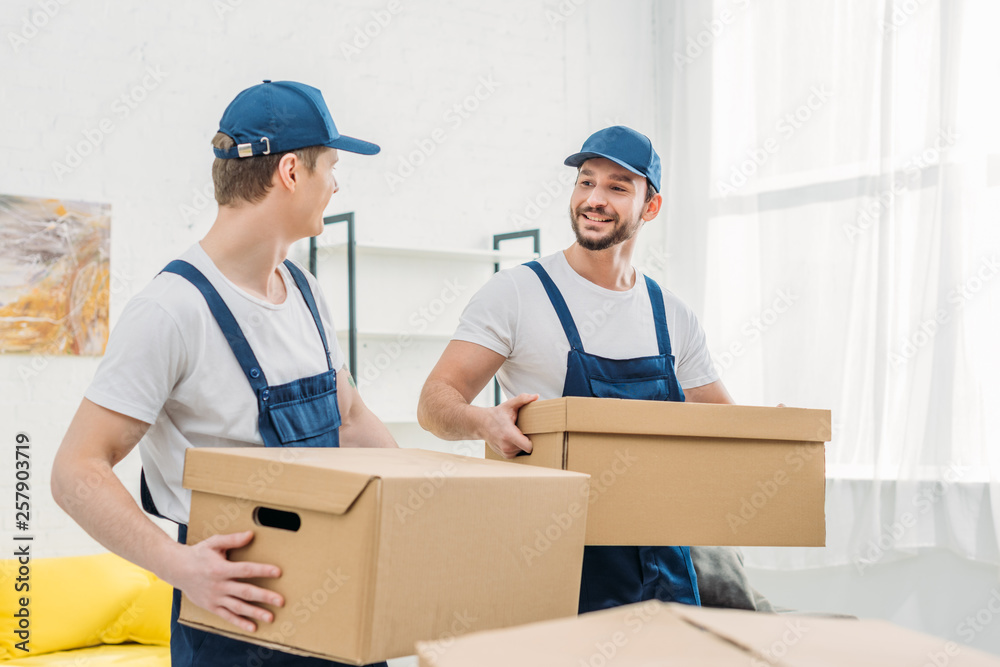 two smiling movers looking at each other while transporting cardboard boxes in apartment two smiling movers looking at each other while transporting cardboard boxes in apartment