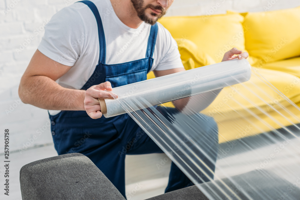 Φωτογραφία cropped view of mover wrapping furniture with roll of stretch film in apartment