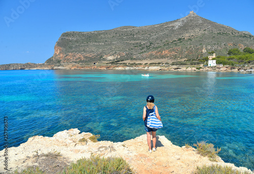  woman on the top of the rock cliff looks at paradise clear torquoise blue water with boats and cloudy blue sky in background in Favignana island, Cala Rossa Beach, Sicily South Italy.