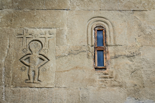 The wall of the church with a small window. An image of an alien or person with crosses, embossed in stone. Georgia, Mtskheta. Religion, Christianity, Orthodoxy.