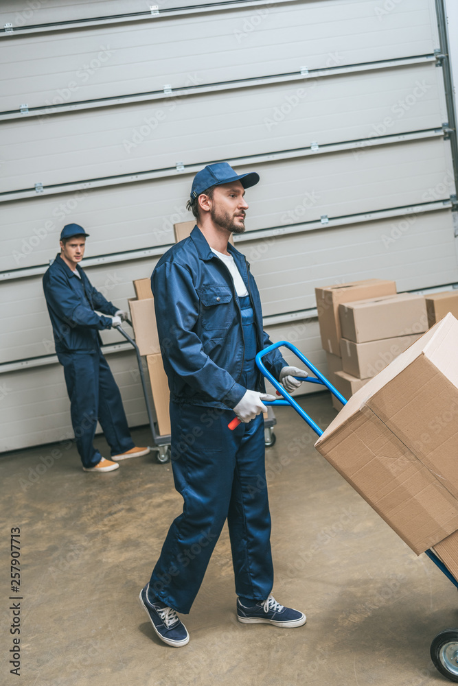 two movers in uniform transporting cardboard boxes with hand trucks in warehouse