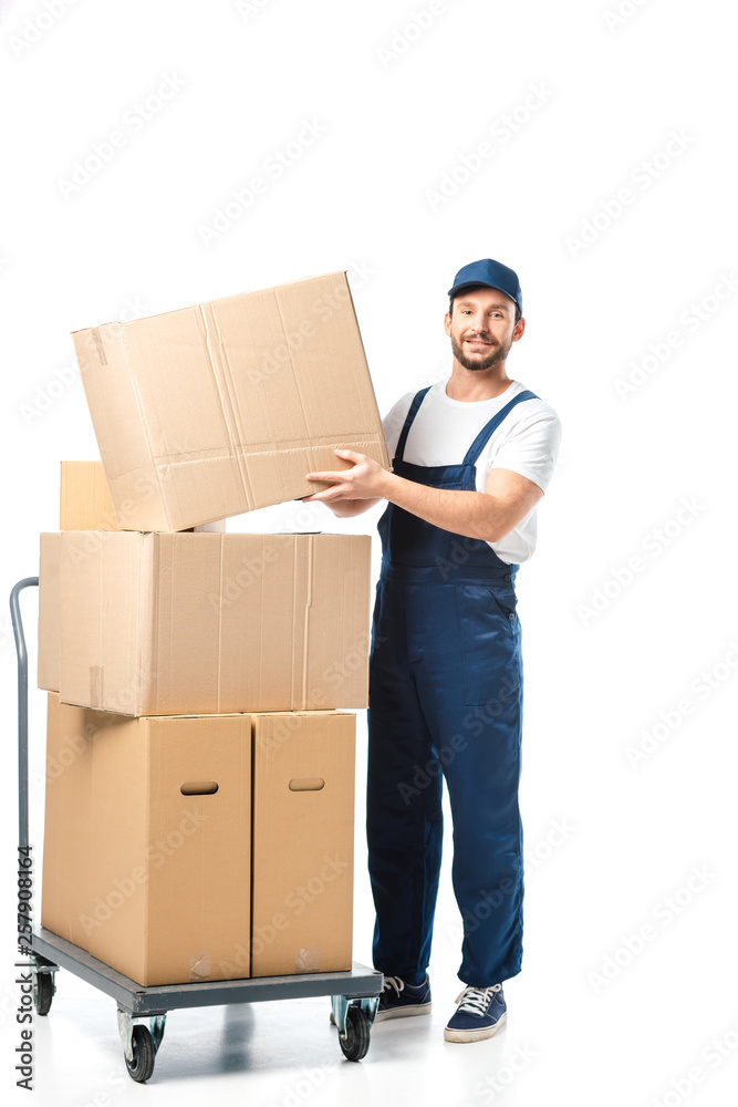 handsome mover in uniform transporting cardboard box near hand truck with packages isolated on white handsome mover in uniform transporting cardboard box near hand truck with packages isolated on white