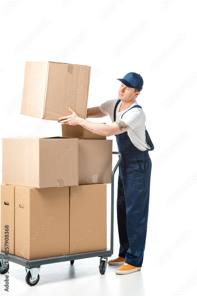 handsome mover in uniform transporting cardboard box near hand truck with packages isolated on white handsome mover in uniform transporting cardboard box near hand truck with packages isolated on white