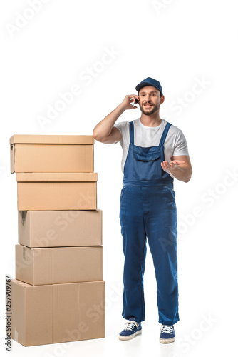 handsome smiling mover with cardboard boxes talking on smartphone and gesturing with hand isolated on white