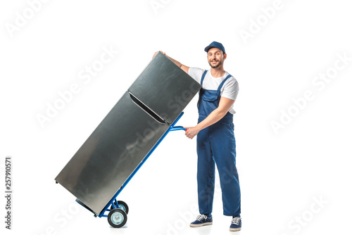 handsome mover in uniform looking at camera and transporting refrigerator on hand truck isolated on white