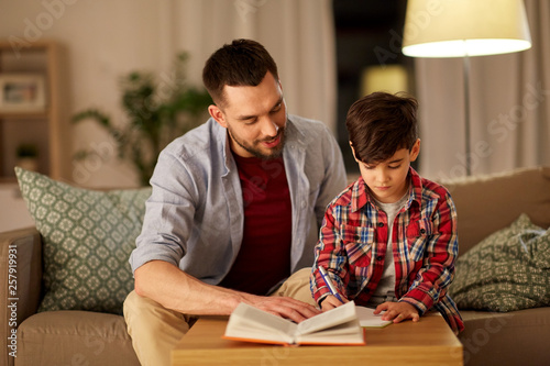 education, family and homework concept - happy father and son with book writing to notebook at home in evening