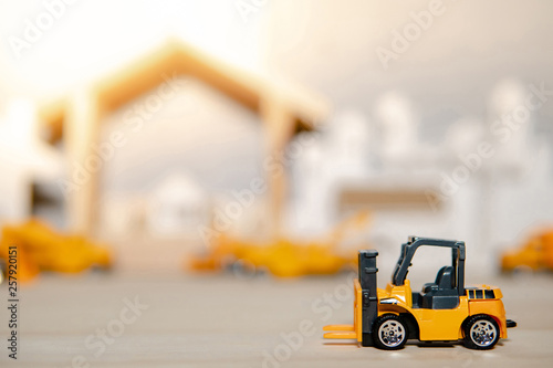 Miniature yellow forklift model on wooden table with blurred house frame and city in the background. Warehouse logistics business or construction industry concept