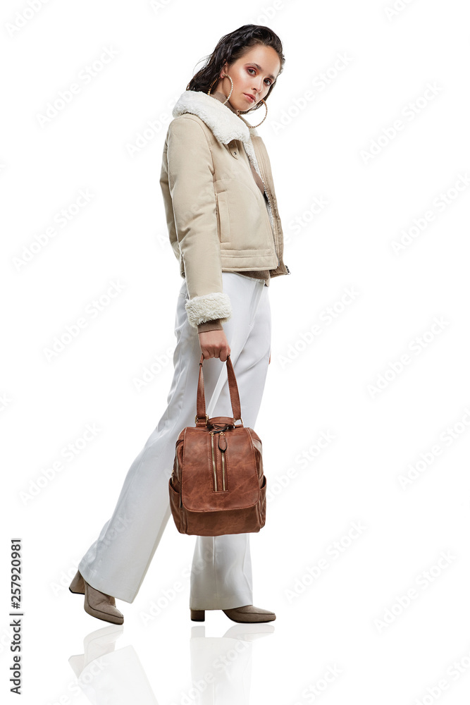 Full length side shot of lady, wearing short Afghan coat and white trousers. The girl is making a step, tilting head and holding shoulder strap of brown rucksack with flap top, adorned with zippers. Full length side shot of lady, wearing short Afghan coat and white trousers. The girl is making a step, tilting head and holding shoulder strap of brown rucksack with flap top, adorned with zippers.