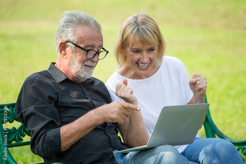 Happy senior loving couple relaxing with laptop computer at park excited together in morning time. old people sitting on a bench in the summer park . Elderly resting .mature relationships. family