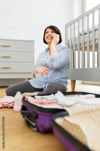 pregnancy, communication and nursery concept - happy pregnant middle-aged woman packing bag or suitcase for maternity hospital and calling on smartphone at home