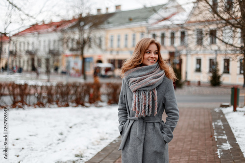 Urban pretty young woman with a beautiful smile in a gray stylish coat in a knitted warm scarf walks around the city near the vintage buildings on a winter day. Attractive happy girl on vacation. photo