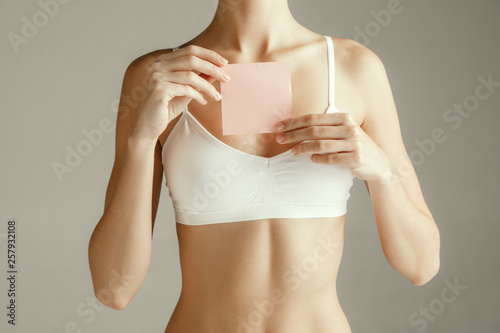 Woman health. Female model holding empty card near breast. Young adult girl with paper for sign or symbol isolated on gray studio background. Cut out part of body. Medical problem and solution.