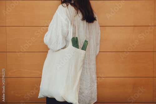 Crop picture of a young woman holding blank textile grocery shopper bag with leek and fresh vegetables inside, wooden wall on the background, concept of healthy farm sustainable shopping