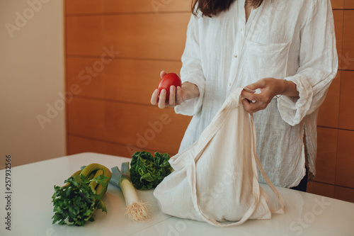 Hipster girl putting greens and fresh leek on the kitchen table out of the cotton reusable tote bag, using eco shopper instead of a plastic bag, concept of healthy lifestyle and saving the planet