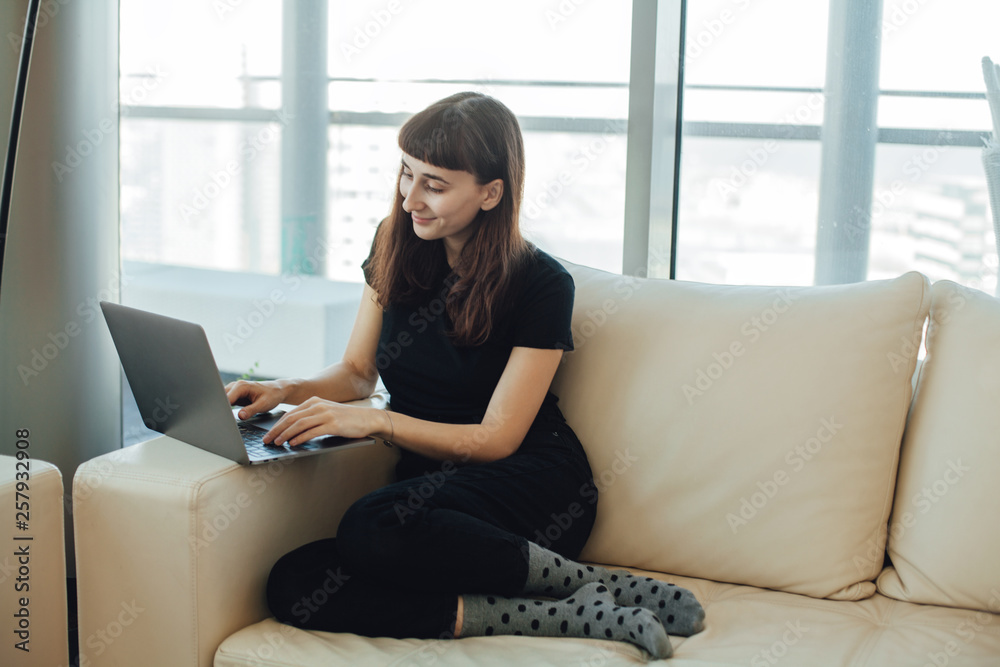 Happy smiling hipster girl typing on a laptop computer keyboard, chatting with family or friends via online messenger while sitting on a cozy sofa ot home with modern interior design Happy smiling hipster girl typing on a laptop computer keyboard, chatting with family or friends via online messenger while sitting on a cozy sofa ot home with modern interior design