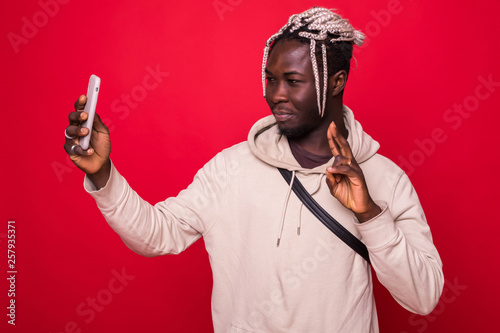 Portrait of happy young african man posing and take a selfie by phone showing peace gesture isolated over red background