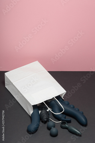A paper shopping bag filled with black silicone sex toys on a pink background. Sex gadget and masturbation device: anal butt plugs, vibrators, vaginal balls, dildo. View from above. Place for text.