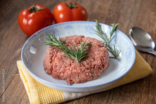 Fresh raw minced meat with rosemary on a plate and tomato on wooden table
