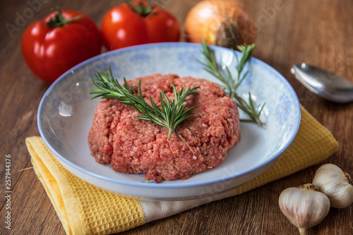 Fresh raw minced meat with rosemary on a plate and tomato, garlic and onion on wooden table