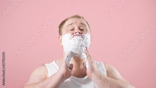 Cheerful happy man with shaving foam on his face doing morning routine procedure isolated over pink background. photo