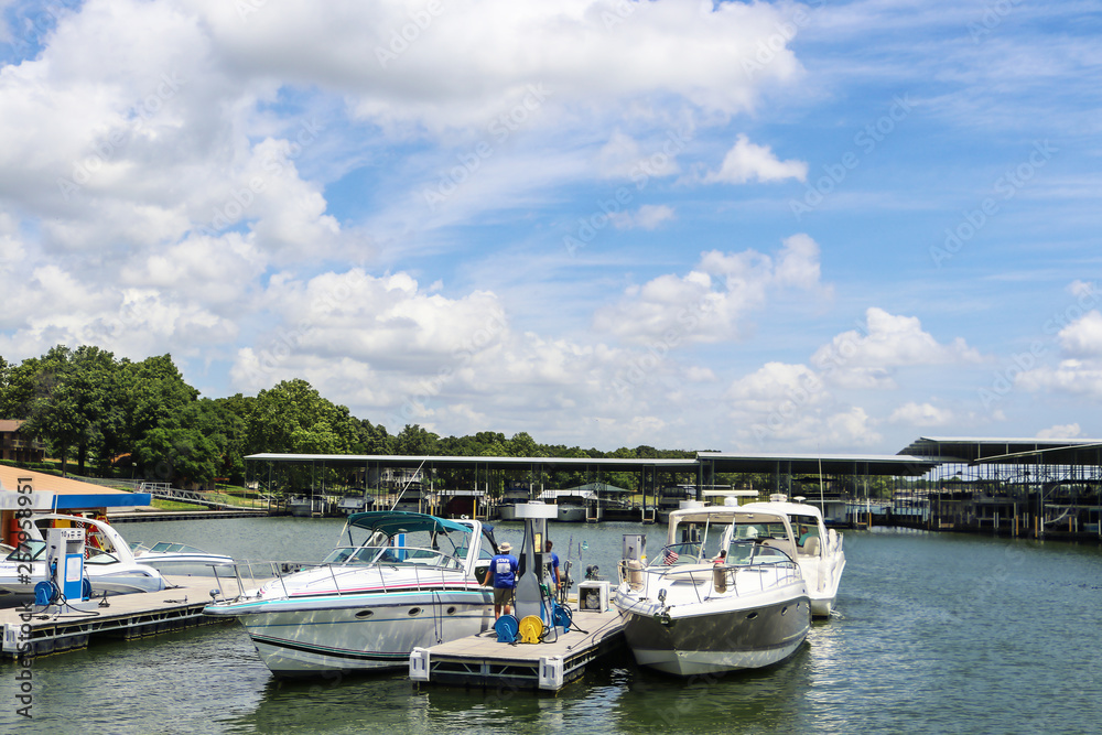 Luxury speedboats fueling up at gas pump at marina on lake with docks and boats behind under beautiful blue cloudy sky