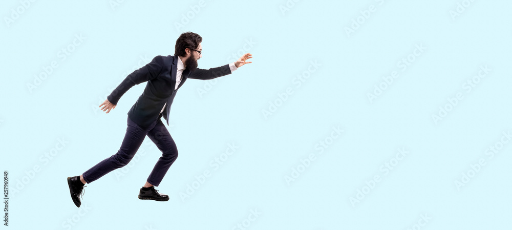 full length portrait of bearded, trendy businessman running against blue background, man pulls hand forward to catch something, panoramic image