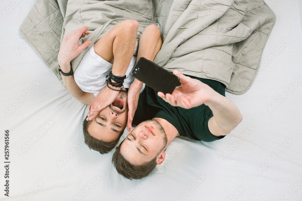 Young gay couple lying under blanket on bed in light room an making selfie on smartphone. Gay couple concept, homosexual