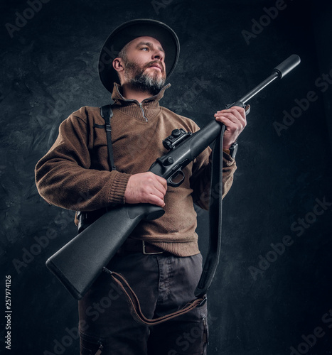 Portrait of a bearded hunter holding a rifle and looking sideways. Studio photo against a dark wall background © Fxquadro
