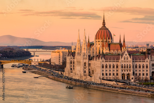 Cityscape of Budapest with bright parliament illuminated by last sunshine before sundown and Danube river with bridge. Pink and purple colors of sky reflecting in water during sunset.