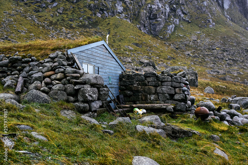 Fotografie Remote blue wooden cabin Stokkvika Hytta at the coast of Lofoten Islands in Norway surrounded by stones, grass and mountains