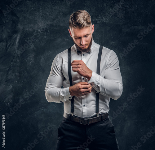 Old-fashioned young man with stylish beard and hair in shirt with bow tie and suspenders buttoning up his sleeve