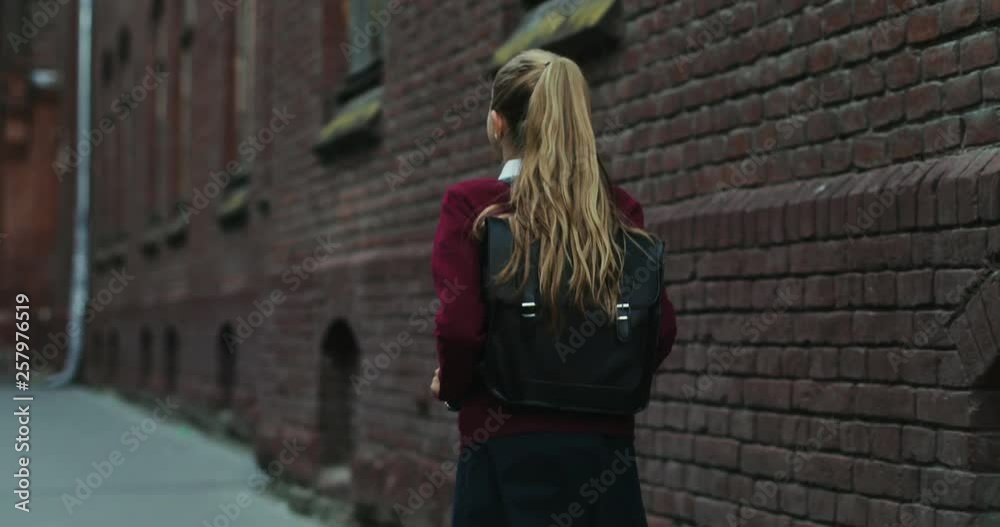 Back view on the Caucasian schoolgirl in the uniform and with a bag walking the street after classes at school. Close up. Rear. Back view on the Caucasian schoolgirl in the uniform and with a bag walking the street after classes at school. Close up. Rear.