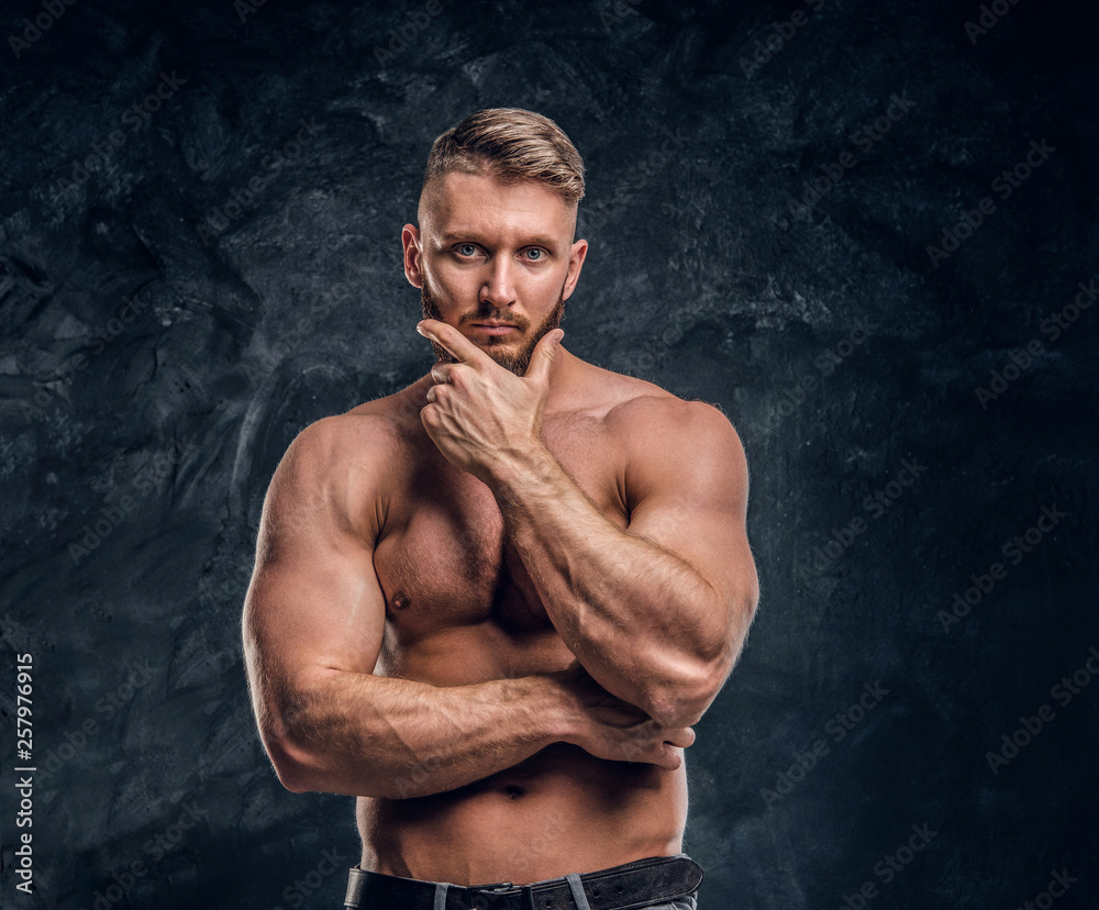 Bearded shirtless male with muscular body posing with hand on chin. Studio photo against dark wall background