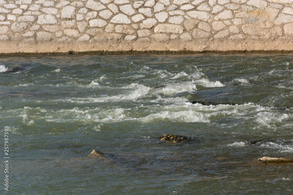 Fototapeta premium Big green river rapids and rock quay on a spring sunny day in city park. Natural background concept. Close up, selective focus
